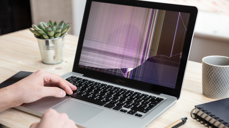 A closeup of a MacBook with a broken screen as it sits on a table next to some desk items, including a notebook, coffee cup, and potted plant