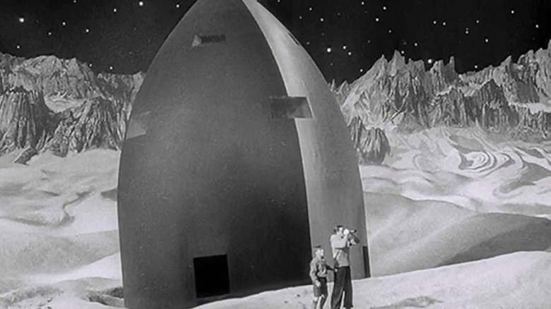 A black and white shot shows two actors surveying the moon's sandy, mountainous terrain through a pair of binoculars in front of their rocket.