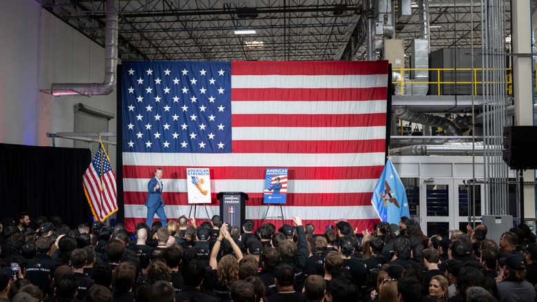 Pete Hegseth gives a thumbs up on stage as he walks to the podium before a large American flag and a crowded audience during his Arsenal of Freedom tour.