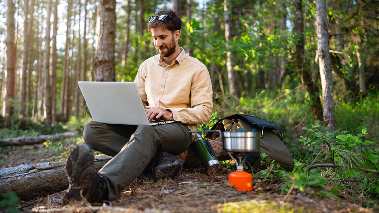 A man using a laptop at a campsite in the woods beside a camping stove and backpack.