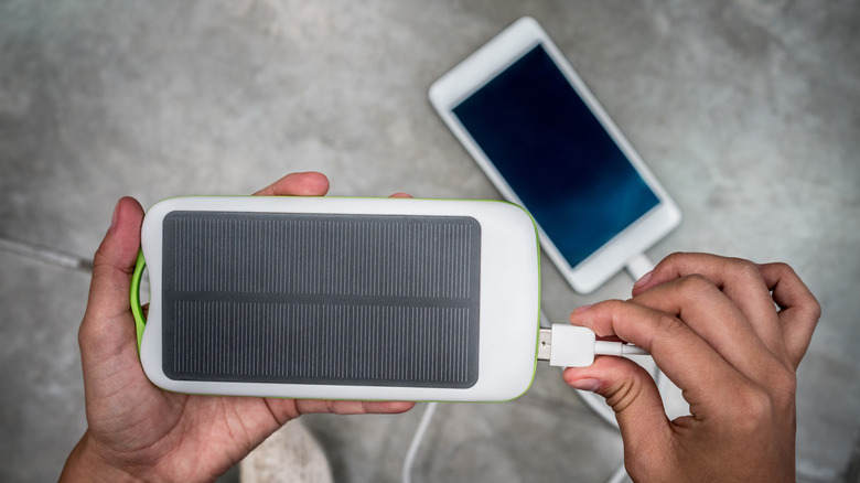 Hands plugging a charging cable into a solar power bank with a smartphone in the background.