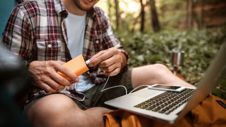 A person connecting a charging cable to a power bank at a campsite beside a laptop and smartphone.