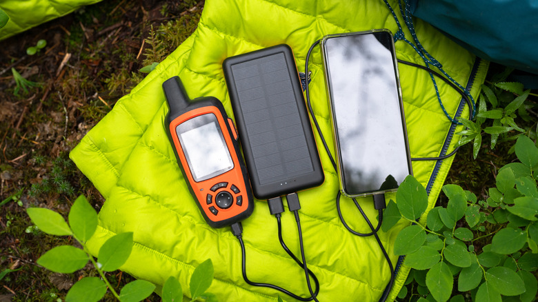 Gadgets, including a smartphone, charging on a solar power bank on a green sleeping bag outdoors.
