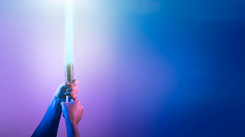 A pair of hands holding up a lightsaber against a background with a purple-to-blue gradient