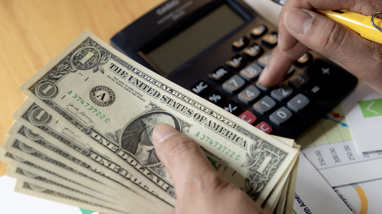 A person holding dollar bills in one hand and a calculator in the other