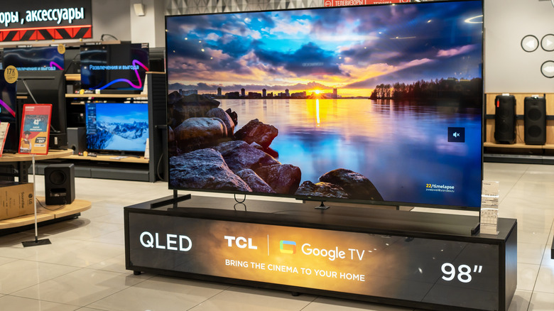A photograph of a TCL 98-inch QLED TV on a showroom floor.