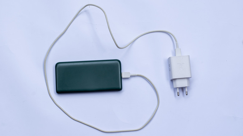 A power bank with a charging cable and adapter plugged in, sitting on a white table
