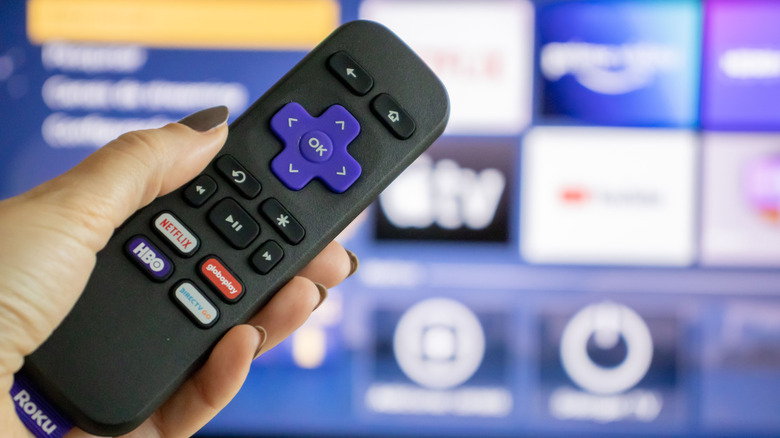A closeup of a hand holding a Roku remote with a blurry TV in the background displaying the Roku Home screen