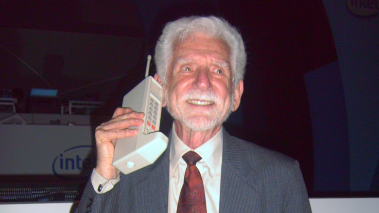Dr. Martin Cooper smiling while holding an old Motorola DynaTAC brick phone.