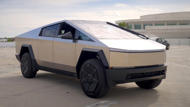 A Tesla Cybertruck with its silver stainless steel exterior sits in a parking lot in front of a concrete building in the background