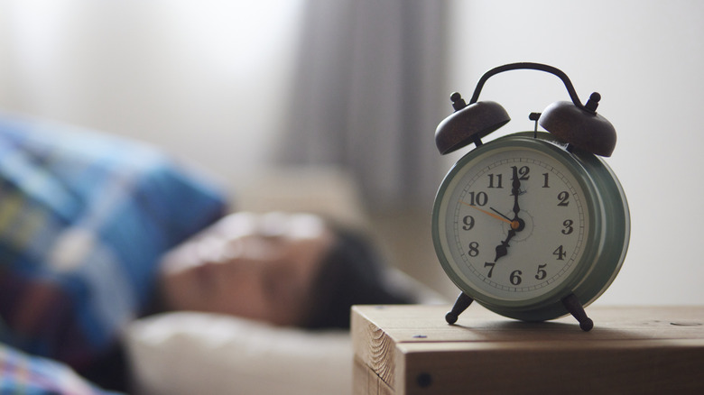 A traditional physical alarm clock on a nightstand with a person in a bed in the background