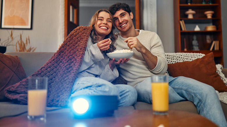 Couple sharing a blanket on a couch with a projector on the table in front of them