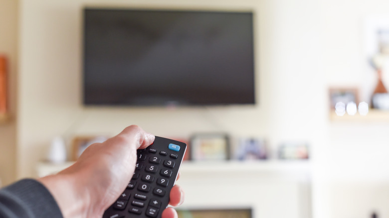 A remote in hand held up to a TV in the background that's off.