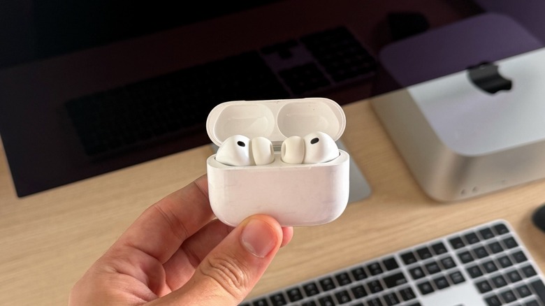 AirPods Pro 3 inside the charging case. In the back, a Mac Studio with the Studio Display
