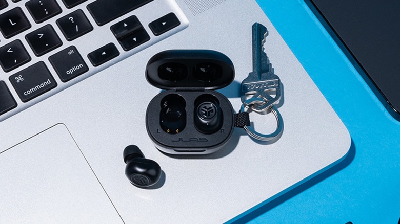 The charging case for the JLabsJBuds Mini Bluetooth Earbuds sits atop a MacBook, one of the earbuds is laying on the laptop, whereas the other is still sitting in the black case