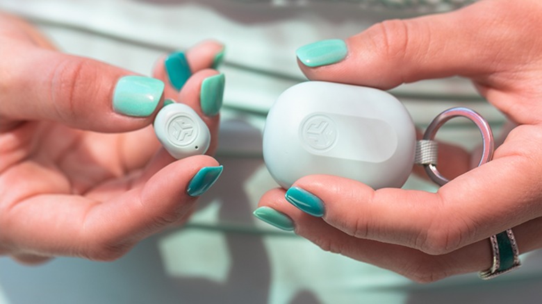 An individual with mint nail polish holds a mint-colored JLabs JBuds mini charging case in one hand and one of the earbuds in the other, the individual is also wearing mint-colored clothes