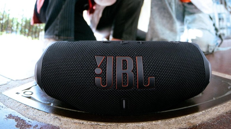 The JBL Charge 6 placed on top of a metal grate in an outdoor setting. A group of people stand in the background.