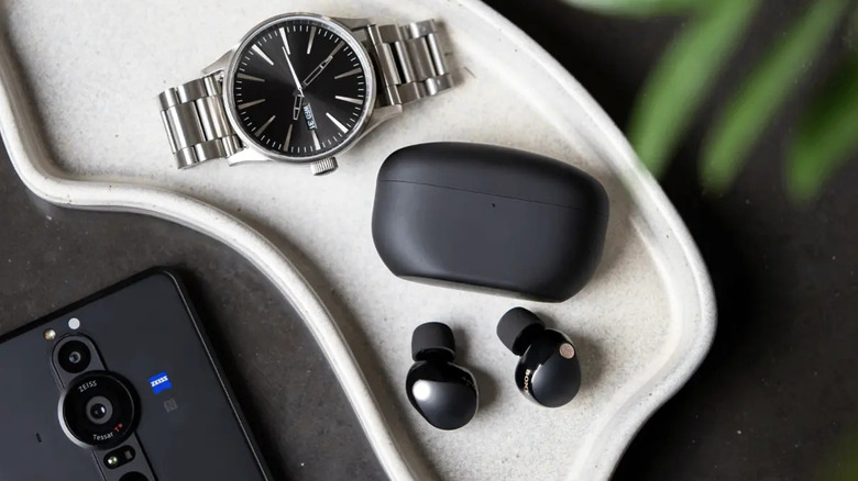 Sony WF-1000XM5 earbuds sittings next to carrying case, a wristwatch, and a smartphone