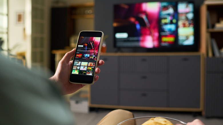 A hand holding a phone showing streaming apps in front of a TV