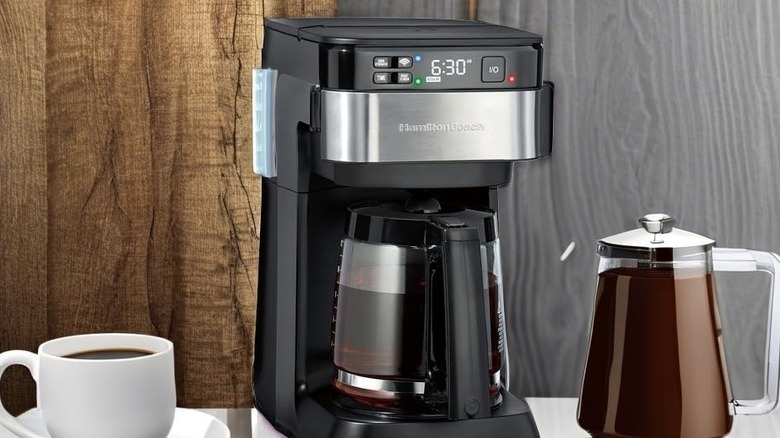 The Hamilton Beach Smart Coffee Maker sitting next to a mug of coffee and a coffee-filled pitcher.