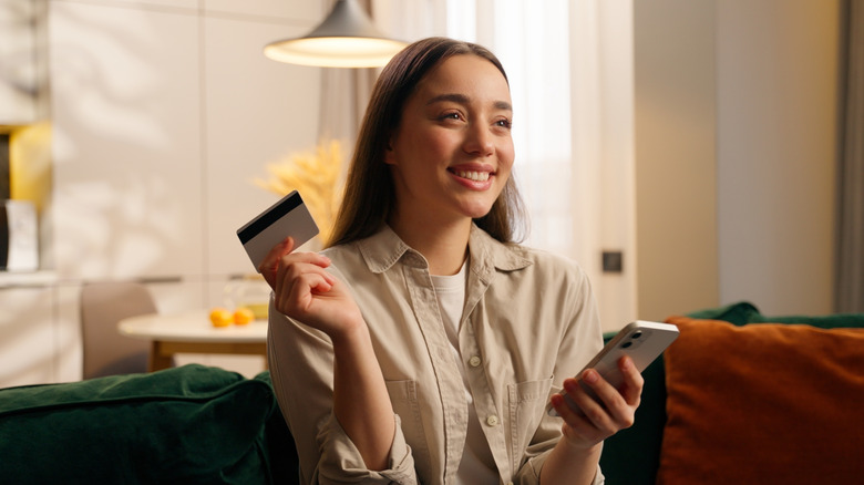 A woman sits on her couch holding her phone in one hand and her credit card in another.