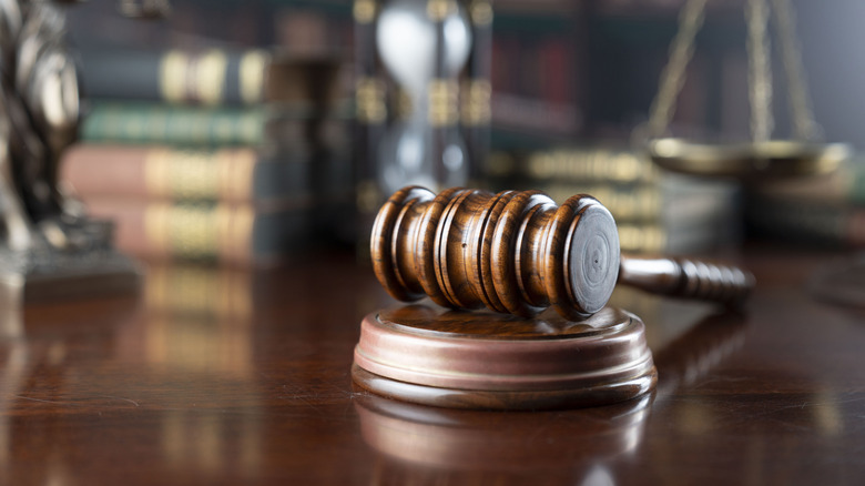 A wooden gavel rests on a dark wood table, books and scales are in the blurred background.