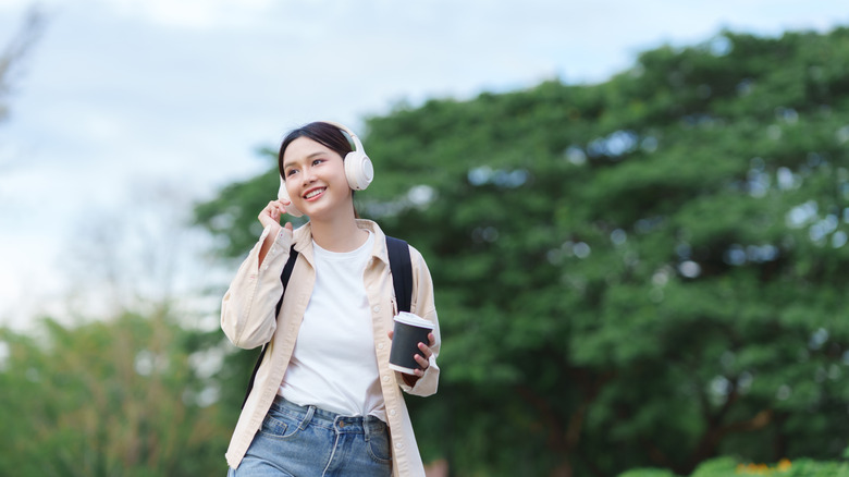 A woman wearing headphones and holding coffee walking outside