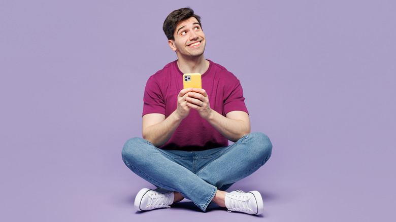 A smiling man holding a phone. He sits on a floor and is against a purple backdrop.