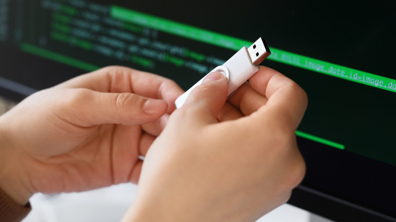 Hands holding a nondescript USB drive in the foreground with code on a screen in the background.