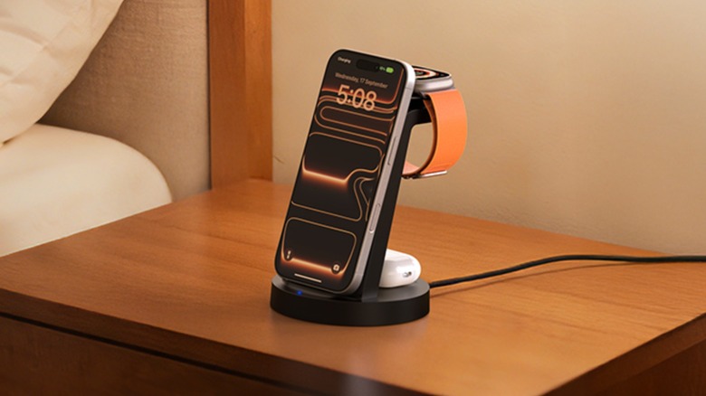 The Anlmz 3-in-1 charging station sitting on a brown dresser next to a bed, the device is charging an iPhone, an Apple Watch, and a pair of AirPods