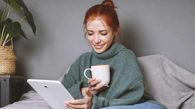 A woman is sitting on the couch with a coffee mug in one hand, a tablet on the other.