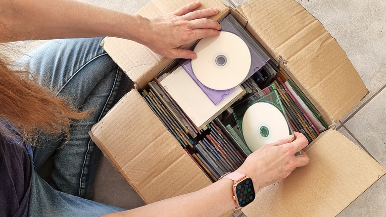 An individual sitting in front of a box of discs, their right hand grabbing a disc in a jewel case while their left hands lays on top of another one
