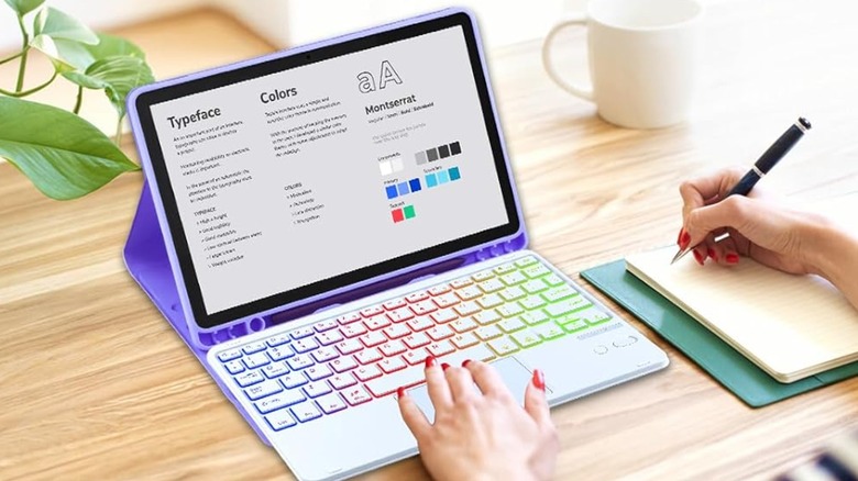 Two hands are on a desk, one hand is hovering over the purple MePadKey Trackpad Case with an Amazon Fire Tablet attached, whereas the right hand is holding a pen and readying to write something on a notepad