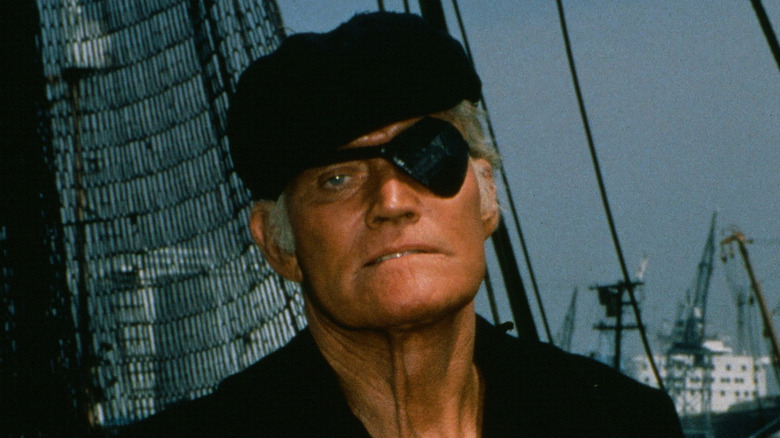 Janos Skorzeny with an eye patch standing on a dock in front of multiple ships.