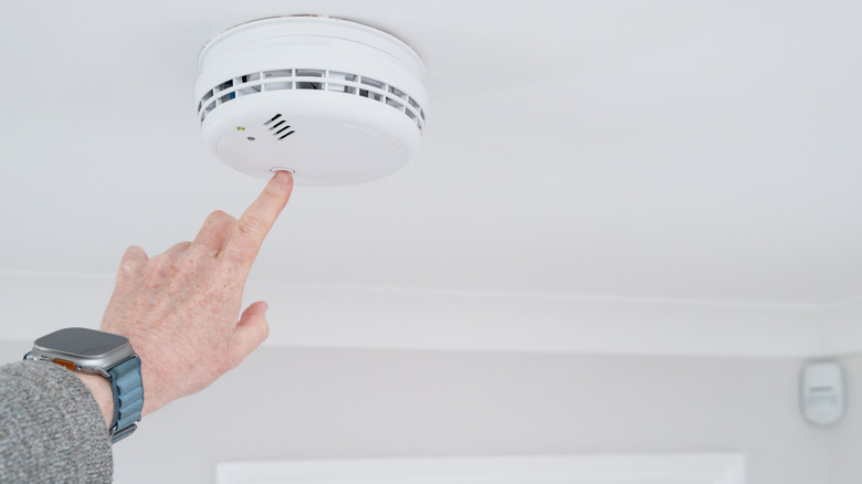 A hand earing a smart watch presses the white reset button of a carbon monoxide detector set on a plain white wall.