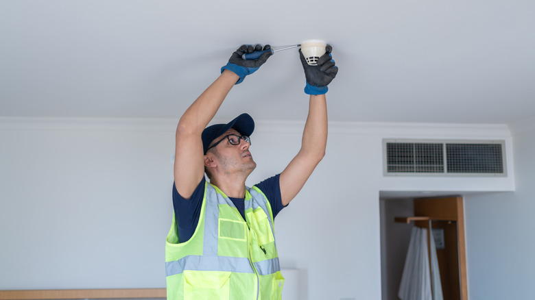A handyman wearing a bright neon vest installs a carbon monoxide detector on a ceiling.