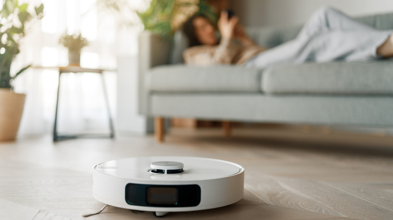 A white robot vacuum cleaning a wood floor while a person in the background lies on a couch checking their phone