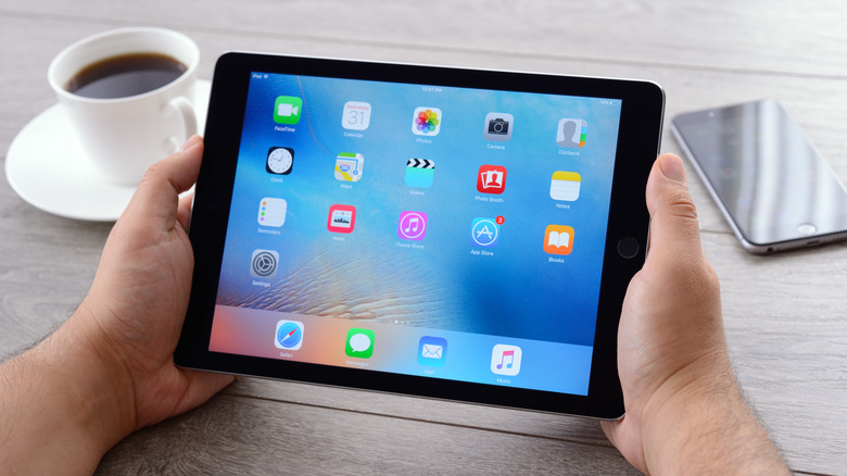 A person holding an Apple iPad, with a cup of coffee and an iPhone on a table behind it