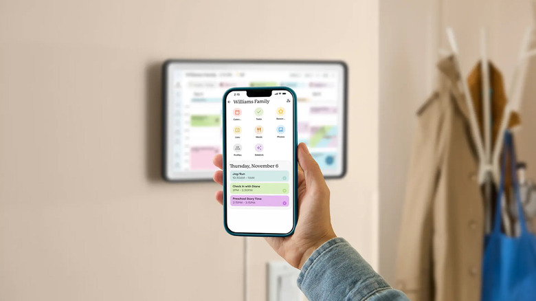A close-up of a phone showing the Skylight app with a Skylight Calendar mounted on the wall in the background.