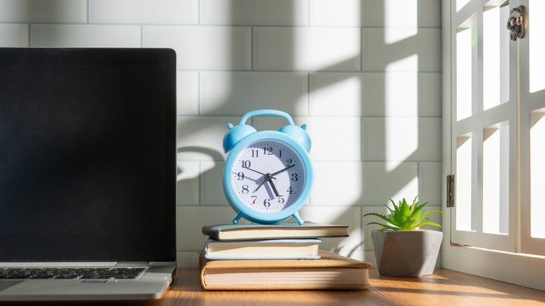 An alarm clock on a stack of books next to a laptop and a window in a white room