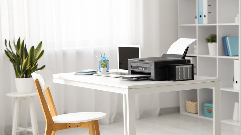 Office setting with a printer resting next to a laptop on a table.