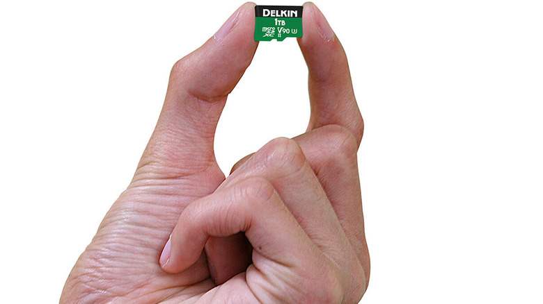 A hand holding a 1TB Delkin MicroSDXC card on a white background