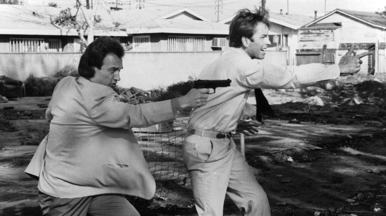 Jim Belushi and John Ritter filming "Real Men." Belushi is pointing a gun, while Ritter aims finger guns