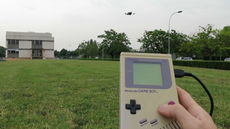 Person holding a Game Boy in a grassy area while a drone hovers nearby
