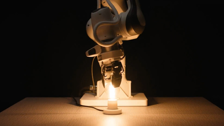 A white Eka robot screws in a lightbulb, causing it to light up a table before a black background