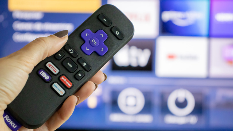 Roku remote in a person's hand with streaming OS on TV in background.