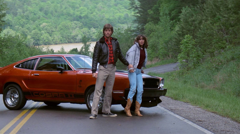 Starman and Jenny standing on the road in front of a 1977 Ford Mustang Cobra II