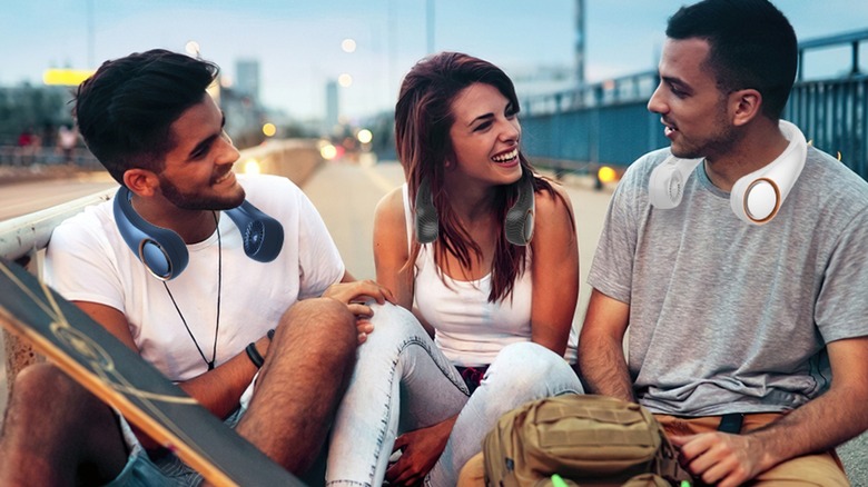 Three individuals smiling together as they sit on an outdoor curb, each is wearing a Penkou Portable Neck Fan