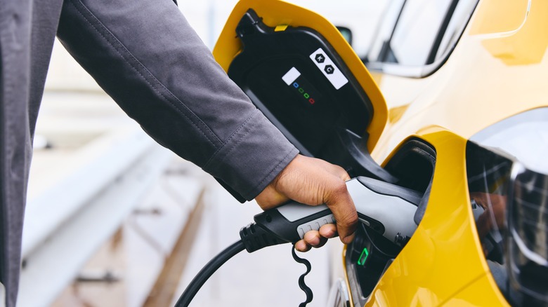 A faceless man wearing a grey jacket plugs an EV charger into a bright yellow car.