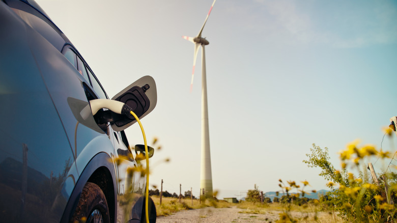 An electric car charges at the end of a dirt road which runs beneath a large windmill.